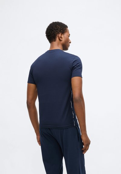 Vīrietis tumši zila īsām piedurknēm krekliņā un biksēs, stāv ar muguru pret vienkāršu baltu fonu.