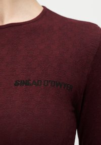 Κοντινό πλάνο πουκαμίσου από ύφασμα βουργουνδίας με διακριτικό μοτίβο κρανίου και σταυρωμένων οστών και μαύρο κείμενο "Sinead O'Dwyer" στο στήθος.