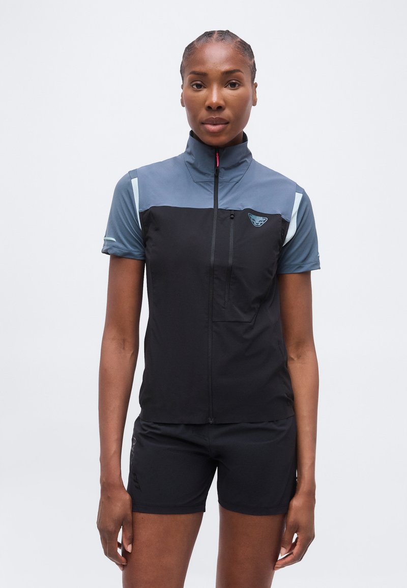 Γυναίκα που φοράει μαύρο και γκρι αμάνικο γιλέκο με φερμουάρ πάνω από μπλε κοντομάνικο μπλουζάκι και μαύρα σορτς, στέκεται μπροστά σε λευκό φόντο.