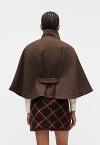 Άτομο με πλεξούδ ες, φοράει καφέ κάπα με ψηλό γιακά και κόκκινη και μαύρη καρό φούστα, με την πλάτη γυρισμένη σε ένα απλό φόντο.