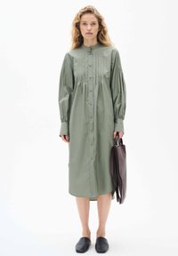 Olajzöld pamut, gombos ruha redőkkel, hosszú ujjakkal és mandarin gallérral, sötétbarna táskával és slip-on cipőkkel párosítva.