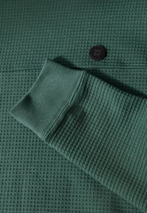 Grüner, strukturiertes Strickgewebe mit Waffelstruktur, das mit einem schwarzen Knopfdetail und einem gerippten Bündchen versehen ist.