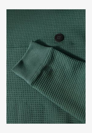 Grüner, strukturiertes Strickgewebe mit Waffelstruktur, das mit einem schwarzen Knopfdetail und einem gerippten Bündchen versehen ist.