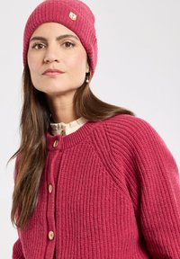 Gilet en tricot et bonnet dans une couleur rose profond. Le gilet présente une texture côtelée, une fermeture à boutons et un col rond. Le bonnet comporte un petit logo.