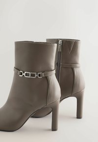 Bottines en cuir gris avec un talon haut, ornées d'une chaîne argentée et d'une fermeture éclair sur le côté. Texture lisse et design structuré.