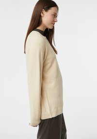 Beżowy sweter o fakturze, z okrągłym dekoltem i ciemnobrązowym prążkowanym kołnierzem. Długie rękawy i luźny krój. Miękki, przytulny materiał.