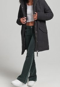 Musta pitkä parka-takki, jossa on turkisvuorattu hupullinen, käytetään valkoisen crop-topin päällä, yhdistettynä vihreisiin corduroy-mekkoihin ja valkoisiin lenkkareihin.