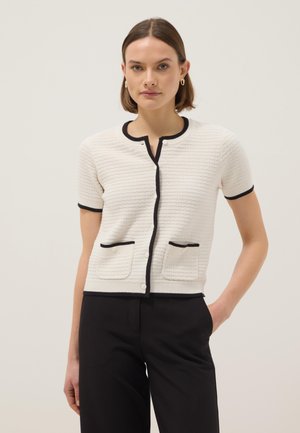 Donna con cardigan bianco texturizzato a maniche corte con bordi neri e tasche, abbinato a pantaloni neri, in piedi davanti a uno sfondo semplice.