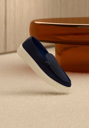 Marineblå slip-on sko med hvit såle, henger over beige teppe, med et blankt brunt rundt bord i det uskarpe bakgrunnen.