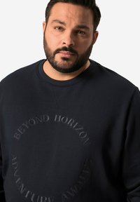 Námořnická mikina se zakulaceným výstřihem, na které se nachází textový grafický prvek se strukturou, znějící "DOBRODRUŽSTVÍ ČEKÁ BEYOND HORIZON." Měkká látka.