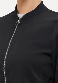 Black bomber jacket with a ribbed collar, silver zipper, and smooth fabric texture. Features a round, close-fitting design with no visible logos.