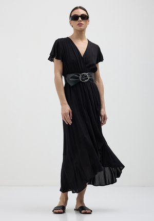 Femme portant une robe noire à enfiler avec une large ceinture, des sandales à glisser noires et des lunettes de soleil rectangulaires foncées, se tenant devant un fond uni.