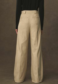 Bredde-beige bukser lavet af tekstureret stof, der har baglommer og høj talje. Designet er minimalistisk med rene linjer.