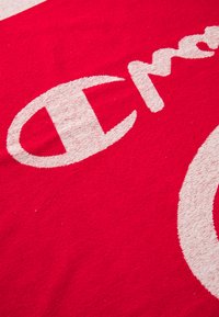 Punainen kangas, jossa on suurikokoinen valkoinen logo pyöristettyine reunoineen, esittää rohkeaa "C"-kirjainta ja tyyliteltyä tekstiä, jolla on pehmeä ja teksturoidun näköinen pinta.