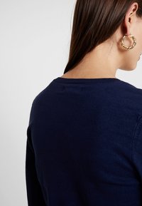 Marinblå stickad tröja med rund halsringning, mjuk textur och lätt detalj vid bakhalsringningen, bärs med stora guldörhängen.