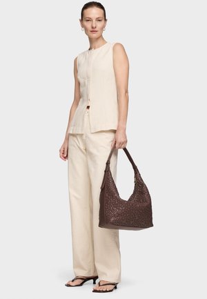 Mujer con blusa sin mangas beige y pantalones, sosteniendo un bolso texturizado marrón oscuro, de pie contra un fondo claro y uniforme.