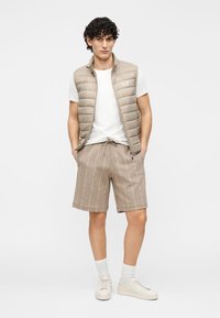 Colete acolchoado bege sobre uma t-shirt branca, combinado com calções bege com padrão subtil. Ténis brancos e meias até ao tornozelo completam o visual.