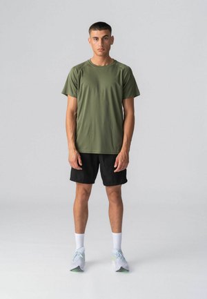 Ung mand står vendt mod fronten iført en olivengrøn kortærmet skjorte, sorte shorts, hvide sokker og hvide løbesko.