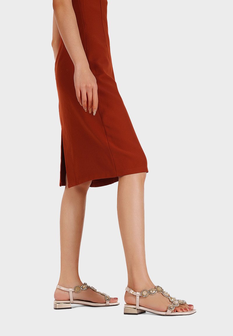 Sandali piatti beige con dettagli decorativi in pietre preziose, presentano un design a fascia e un tacco a blocco basso. Indossati con un vestito aderente color ruggine.