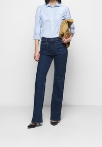 Kvinna som bär en randig blå och vit skjorta, mörkblå hög midja jeans, svarta klackar, håller en vikt sandfärgad väska mot en enkel vit bakgrund.