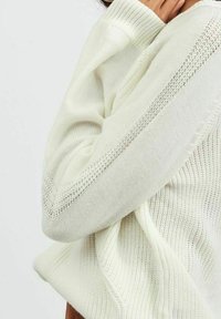 Cream knit sweater with ribbed texture, featuring open-knit accents along the sleeves and hem, showcasing a relaxed fit.