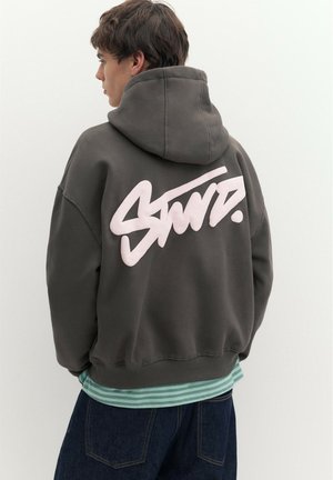 Jeune personne portant un sweat à capuche gris foncé avec de grandes lettres blanches stylisées dans le dos, superposé à une chemise rayée verte et un jean.