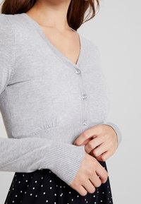 Gray knitted cardigan with a V-neck, three decorative buttons, and ribbed cuffs. Smooth texture with gathered detailing at the waist.