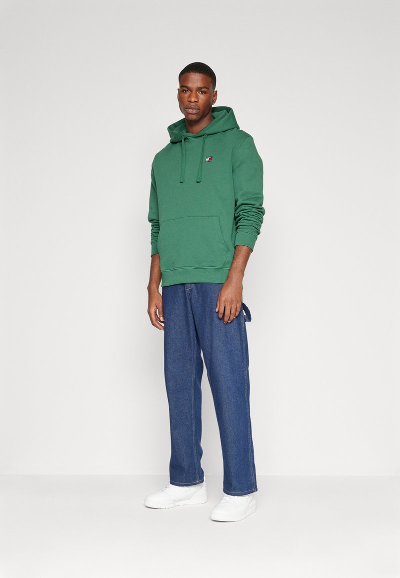 Felpa verde con cappuccio e tasca frontale, con un piccolo logo. Abbinata a pantaloni in denim blu larghi e scarpe da ginnastica bianche.