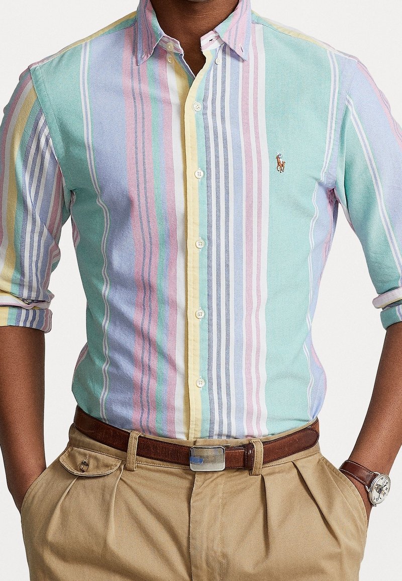 Camisa de botones a rayas con colores pastel (rosa, azul, amarillo) en anchos variados, con un pequeño emblema en el pecho y mangas arremangadas.