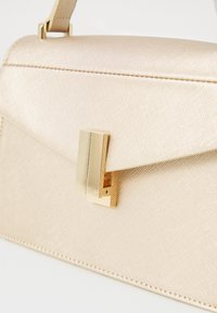Structured handbag in light gold textured material with a magnetic clasp and smooth accents, featuring linear stitching along the edges.