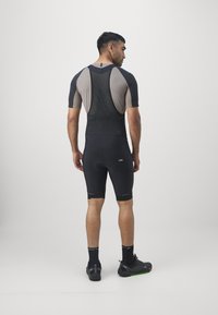 Tenue de cyclisme noire avec un design ajusté, dos en mesh et panneau beige contrastant. Le short arrive jusqu'à mi-cuisse, associé à des chaussures et des chaussettes noires.