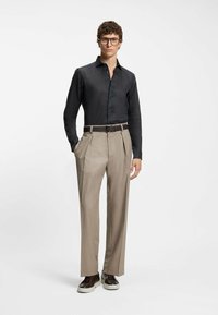 Black long-sleeve shirt paired with beige pleated trousers and brown sneakers. The shirt features a button-up front and a smooth texture.