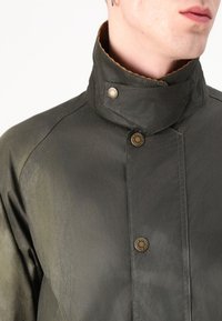 Casaco verde oliva com gola alta, apresentando dois botões de metal e um tecido texturizado. Detalhes de costura notáveis ao longo das costuras.