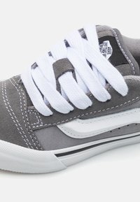 Sneaker en daim gris avec des lacets blancs, comportant un col rembourré, une étiquette de logo noir et blanc, et une semelle blanche en caoutchouc avec des accents noirs.