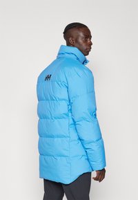 Jachetă puffer de culoare albastră deschisă, cu guler înalt, design matlasat orizontal și logo negru pe spate. Materialul pare neted și lucios.