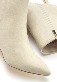 Botas de tobillo beige en tejido texturizado con punta afilada y cremallera lateral con herrajes dorados. Forro suave y detalles de diseño sutiles.