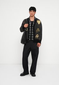 Cardigan negro de punto con gráficos multicolores, superpuesto a una camisa de cuadros. Combinado con pantalones cargo negros y zapatos negros, sosteniendo un bolso negro.