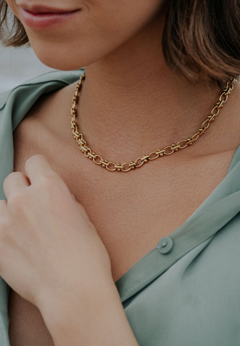 Złoty naszyjnik łańcuchowy z owalnymi ogniwami, z połyskującym wykończeniem. Noszony na tle koloru skóry, uzupełniony przez jasną zieloną koszulę.