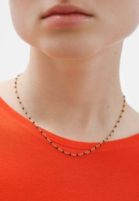 Collar de cadena de oro con cuentas negras, llevado sobre una blusa roja de escote redondo. Superficie suave, diseño delicado con elementos alternos.