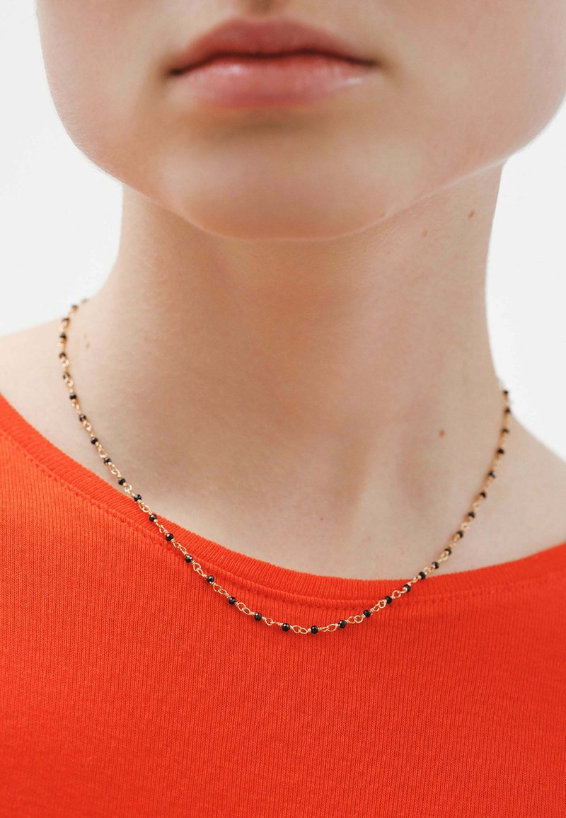 Collar de cadena de oro con cuentas negras, llevado sobre una blusa roja de escote redondo. Superficie suave, diseño delicado con elementos alternos.