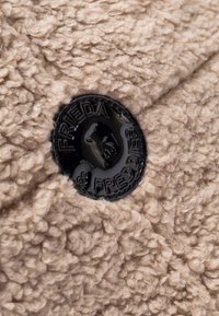 Close-up van een zwarte plastic knop met een verhoogd hondenlogo en de tekst "Frieda & Freddies" op een beige textuur fleece stof.