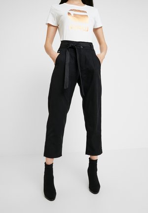 Femme portant un t-shirt blanc avec un imprimé doré, un pantalon noir court taille haute avec ceinture nouée, et des bottines noires, debout les mains dans les poches.