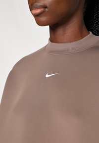 Hnědá mikina s kulatým výstřihem vyrobená z měkké tkaniny, se žebrovaným límečkem a bílým logem Nike na hrudi. Hladký povrch, minimalistický design.