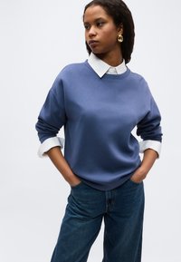 Γυναίκα που φοράει μπλε φούτερ πάνω από λευκό πουκάμισο με γιακά και μανίκια γυρισμένα, συνδυασμένο με μπλε τζιν και χρυσούς κρίκους στα αυτιά, με τα χέρια στις τσέπες.