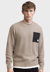 Maglione beige a maglia con un motivo testurizzato, polsini e orlo a coste, e una tasca nera con dettagli del logo sul lato sinistro del petto.