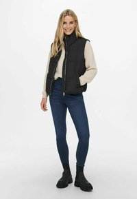 Gilet rembourré noir, fermeture éclair à l'avant, col montant, porté par-dessus un pull léger. Associé à un jean skinny bleu foncé et des bottines noires massives.