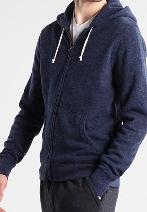 Man draagt een donkerblauwe hoodie met ritssluiting en witte trekkoorden en een wit shirt eronder, met één hand gedeeltelijk in de zak.