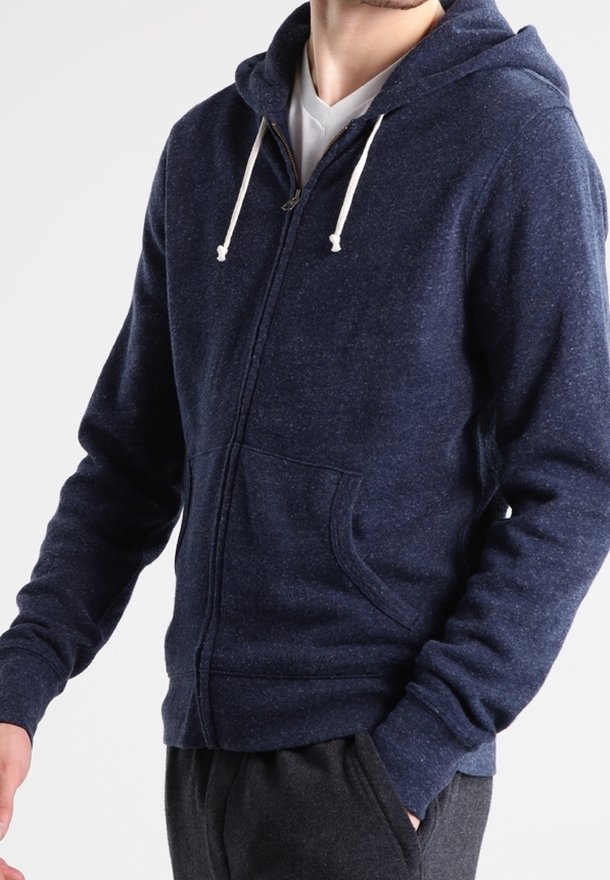 Homme portant un sweat à capuche zippé bleu foncé avec des cordons blancs et une chemise blanche en dessous, avec une main partiellement dans la poche.