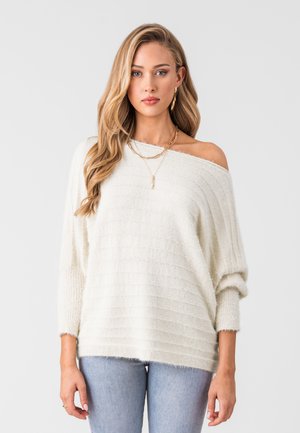 Maglione bianco sciancrato con design a coste testurizzate, maniche lunghe e vestibilità rilassata, indossato sopra jeans azzurri chiari.