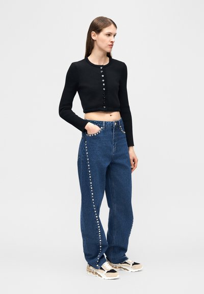 Mujer con cárdigan negro corto con botones y vaqueros de tiro alto azules decorados con tachuelas plateadas y zapatillas blancas beige, de pie de perfil.